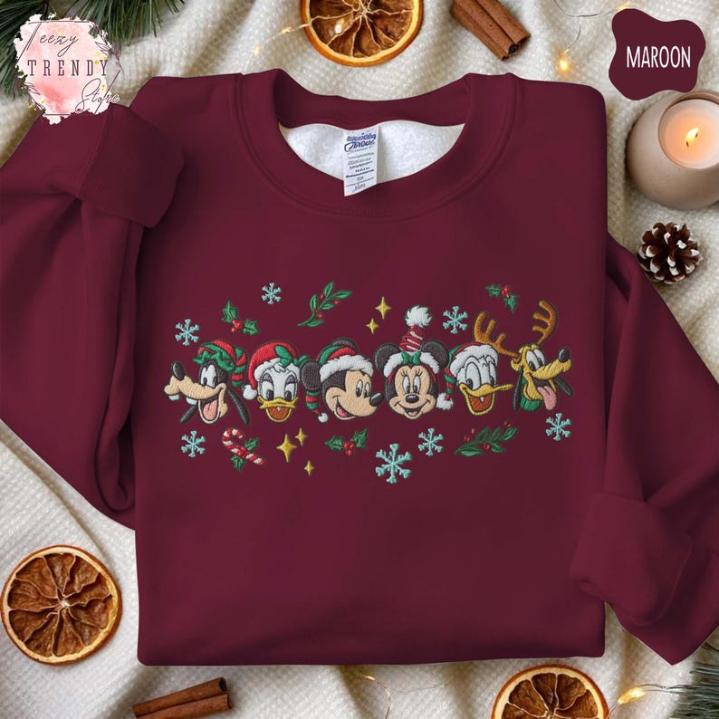 May include: Maroon sweatshirt featuring embroidered Disney characters in festive holiday attire. The design includes Mickey Mouse, Minnie Mouse, Donald Duck, Goofy, and Pluto, with snowflakes and holly accents. A cozy and colorful holiday-themed garment.