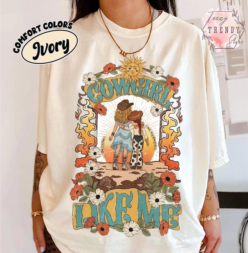 May include: An ivory t-shirt featuring a graphic print of two figures in cowboy hats and boots standing together. The design includes the text COWGIRL LIKE ME, a sun, flowers, and flame-like patterns in orange, yellow, blue, and brown tones.