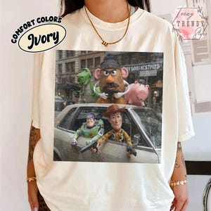 Puede incluir: Una camiseta de color marfil con un estampado gr&aacute;fico de personajes de Toy Story, incluidos Buzz Lightyear, Woody, Rex, Mr. Potato Head y Hamm. Los personajes aparecen en una calle con un coche, cada uno sosteniendo una peque&ntilde;a pistola de juguete negra.