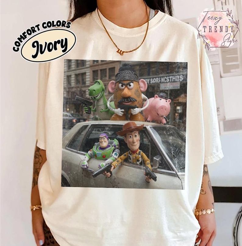 Puede incluir: Una camiseta de color marfil con un estampado gr&aacute;fico de personajes de Toy Story, incluidos Buzz Lightyear, Woody, Rex, Mr. Potato Head y Hamm. Los personajes aparecen en una calle con un coche, cada uno sosteniendo una peque&ntilde;a pistola de juguete negra.