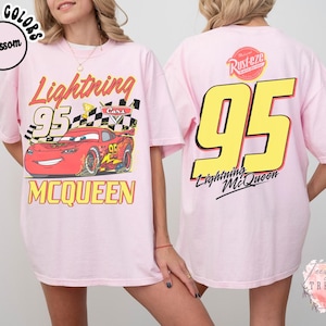 May include: Light pink t-shirt featuring a graphic of Lightning McQueen from the Cars movie. The front of the shirt displays the character with the number 95 and the name McQueen. The back of the shirt has a large yellow 95 and the Lightning McQueen logo.