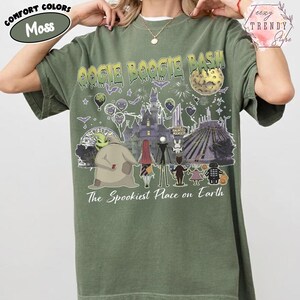 May include: A moss green t-shirt featuring a graphic print with the text OOGIE BOOGIE BASH and The Spookiest Place on Earth. The design includes illustrations of characters from The Nightmare Before Christmas, a castle, a moon, and fireworks.