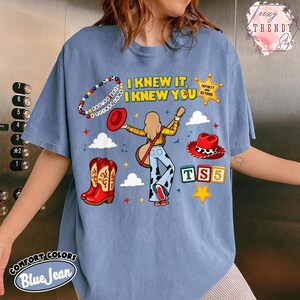 May include: A blue jean coloured t-shirt featuring a graphic of a person with long hair holding a guitar, surrounded by a beaded bracelet, a red hat, a cow print hat, red cowboy boots, a yellow star, and blocks with the text TS5 and I KNEW IT.
