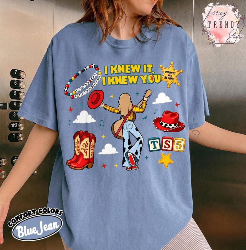 May include: A blue jean coloured t-shirt featuring a graphic of a person with long hair holding a guitar, surrounded by a beaded bracelet, a red hat, a cow print hat, red cowboy boots, a yellow star, and blocks with the text TS5 and I KNEW IT.