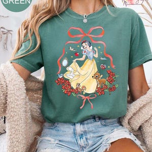 Peut inclure: T-shirt vert clair avec un motif de Blanche-Neige en robe jaune, entour&eacute;e de fleurs rouges, d'oiseaux et d'animaux de la for&ecirc;t. Le t-shirt porte le texte "LIGHT GREEN" dans un cercle en haut &agrave; gauche.
