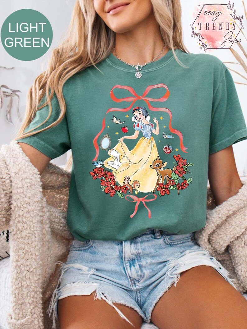 Peut inclure: T-shirt vert clair avec un motif de Blanche-Neige en robe jaune, entour&eacute;e de fleurs rouges, d'oiseaux et d'animaux de la for&ecirc;t. Le t-shirt porte le texte "LIGHT GREEN" dans un cercle en haut &agrave; gauche.
