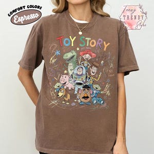 May include: Espresso-colored t-shirt featuring a graphic of Toy Story characters. The design includes Woody, Buzz Lightyear, Rex, and other characters from the animated film. The shirt has the words "Toy Story" in a colorful font.
