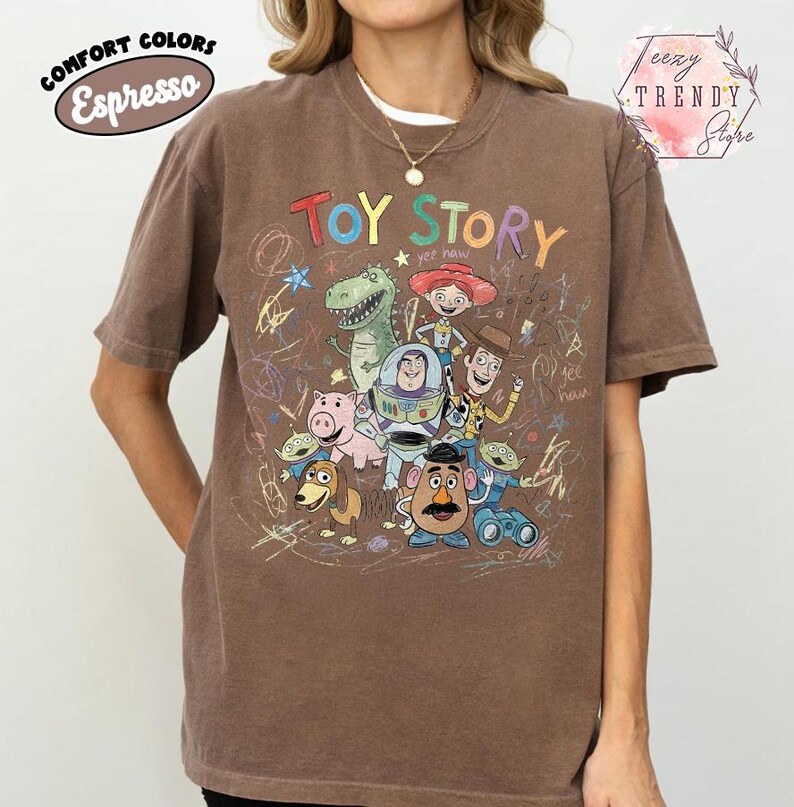 May include: Espresso-colored t-shirt featuring a graphic of Toy Story characters. The design includes Woody, Buzz Lightyear, Rex, and other characters from the animated film. The shirt has the words "Toy Story" in a colorful font.