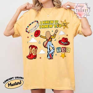 May include: A mustard yellow t-shirt featuring a graphic print with the text I KNEW IT, I KNEW YOU, a beaded bracelet, a sheriff badge, a red hat, a guitar, cowboy boots, and alphabet blocks spelling TS5, surrounded by clouds and stars.