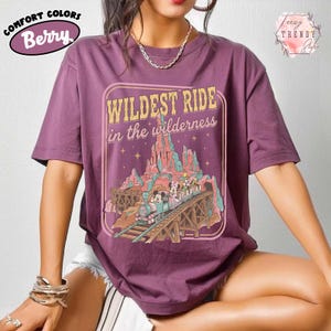 Może przedstawiać: Koszulka Comfort Colors w kolorze jagodowym z grafiką przedstawiającą przejażdżkę kolejką g&oacute;rską z napisem "WILDEST RIDE in the wilderness". Projekt zawiera g&oacute;rski krajobraz i postacie z kresk&oacute;wek. Koszulka ma kr&oacute;tkie rękawy i okrągły dekolt.