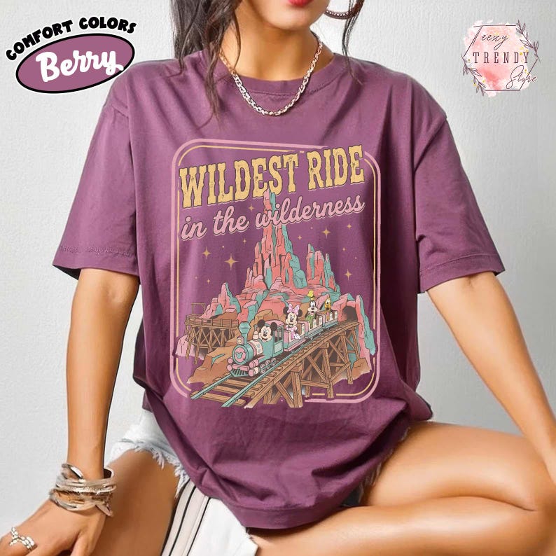 Może przedstawiać: Koszulka Comfort Colors w kolorze jagodowym z grafiką przedstawiającą przejażdżkę kolejką g&oacute;rską z napisem "WILDEST RIDE in the wilderness". Projekt zawiera g&oacute;rski krajobraz i postacie z kresk&oacute;wek. Koszulka ma kr&oacute;tkie rękawy i okrągły dekolt.