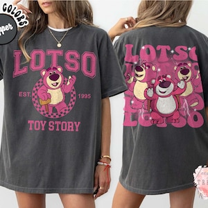 May include: Dark gray t-shirt featuring a pink Lotso bear graphic from Toy Story. The front design includes the word "LOTSO" above the bear, with "EST. 1995" and "TOY STORY" below. The back of the shirt has a larger pink graphic of Lotso and two other bears.