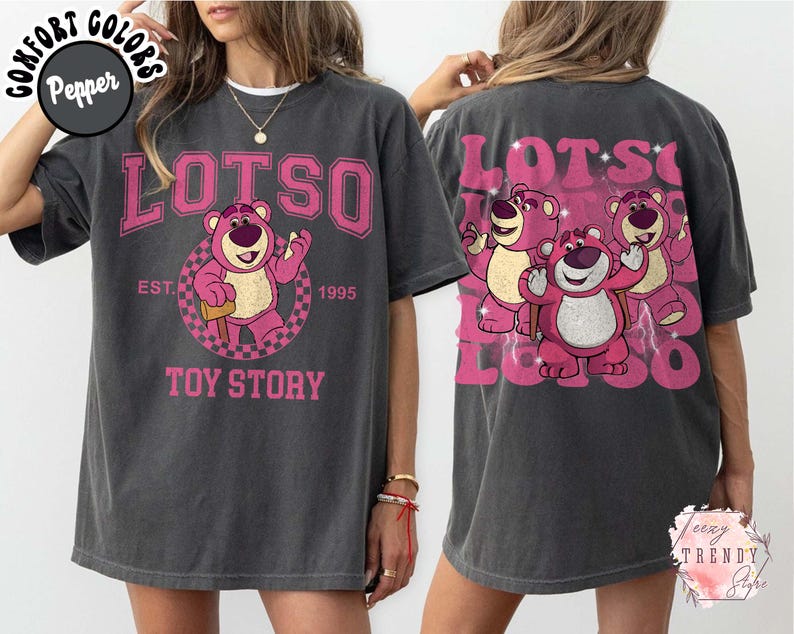 May include: Dark gray t-shirt featuring a pink Lotso bear graphic from Toy Story. The front design includes the word "LOTSO" above the bear, with "EST. 1995" and "TOY STORY" below. The back of the shirt has a larger pink graphic of Lotso and two other bears.