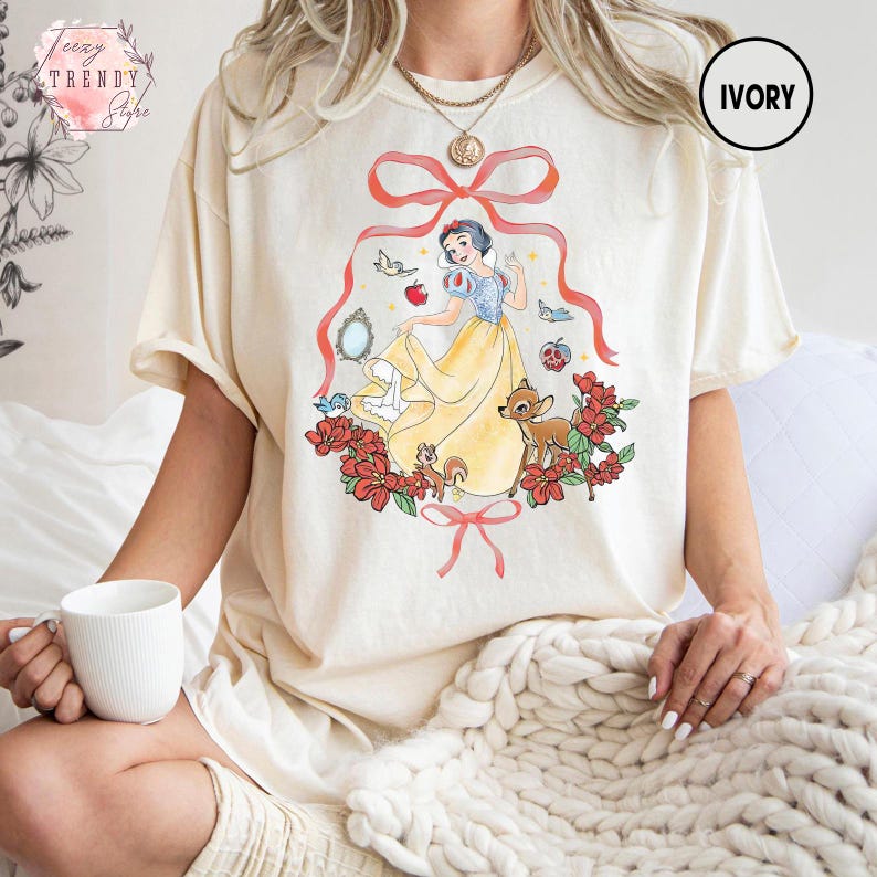 Peut inclure: T-shirt ivoire avec une illustration color&eacute;e de Blanche-Neige. Le motif repr&eacute;sente Blanche-Neige dans une robe jaune, entour&eacute;e de fleurs rouges, d'un cerf et d'autres cr&eacute;atures de la for&ecirc;t. Le texte "IVORY" est dans un cercle en haut &agrave; droite.