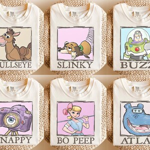 May include: Six cream-coloured t-shirts featuring cartoon characters from the Toy Story franchise. Each shirt has a different character: Bullseye, Slinky, Buzz, Snappy, Bo Peep, and Atlas, each in a framed design with their name below.
