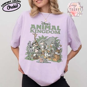 May include: A lilac t-shirt with the text "Disney's Animal Kingdom" in green. The shirt features a graphic of various Disney characters and animals. The shirt also has the text "Comfort Colors Orchid" in a black oval.