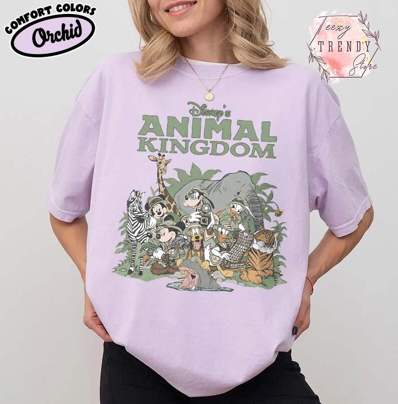 May include: A lilac t-shirt with the text "Disney's Animal Kingdom" in green. The shirt features a graphic of various Disney characters and animals. The shirt also has the text "Comfort Colors Orchid" in a black oval.