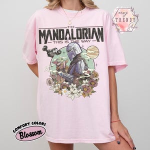 May include: Pale pink t-shirt with a graphic of The Mandalorian and Grogu, set against a floral and planetary backdrop. The text "THE MANDALORIAN - THIS IS THE WAY" is printed across the top. The shirt has a crew neck and short sleeves.
