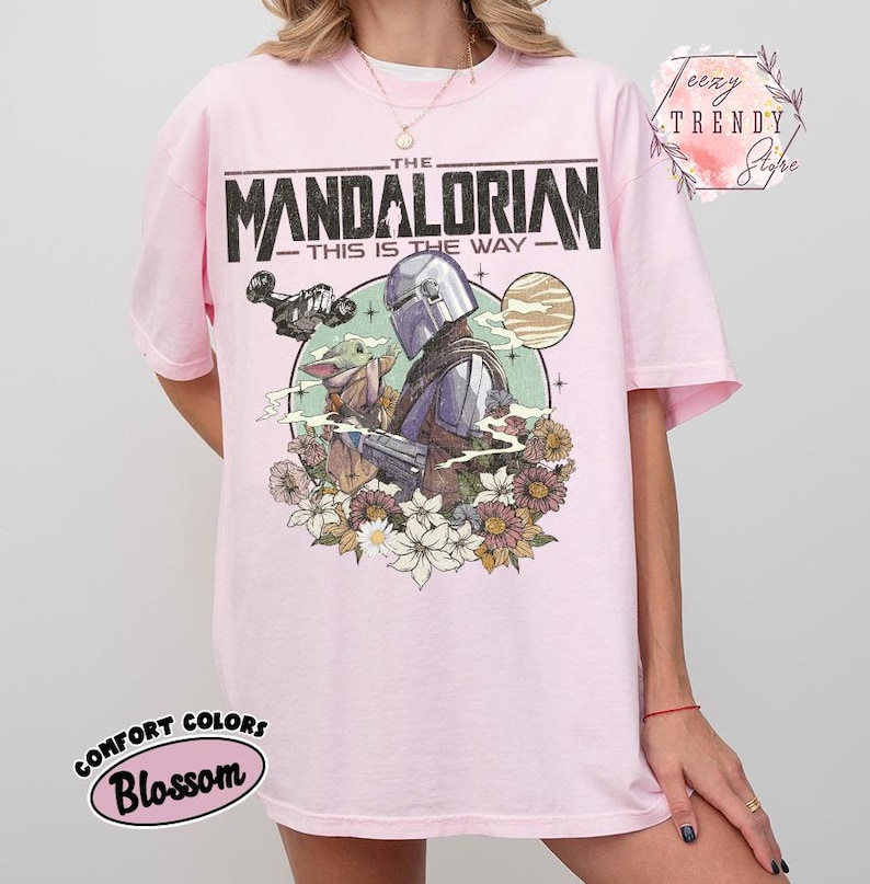 May include: Pale pink t-shirt with a graphic of The Mandalorian and Grogu, set against a floral and planetary backdrop. The text "THE MANDALORIAN - THIS IS THE WAY" is printed across the top. The shirt has a crew neck and short sleeves.