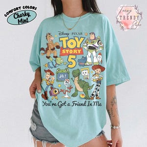May include: A light mint green t-shirt featuring a graphic print of Disney Pixar Toy Story characters including Woody, Buzz Lightyear, Jessie, Rex, and Forky. The text reads Toy Story 5 and You're Got a Friend In Me with a small Hi sign.