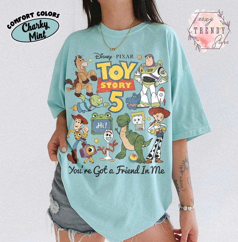 May include: A light mint green t-shirt featuring a graphic print of Disney Pixar Toy Story characters including Woody, Buzz Lightyear, Jessie, Rex, and Forky. The text reads Toy Story 5 and You're Got a Friend In Me with a small Hi sign.