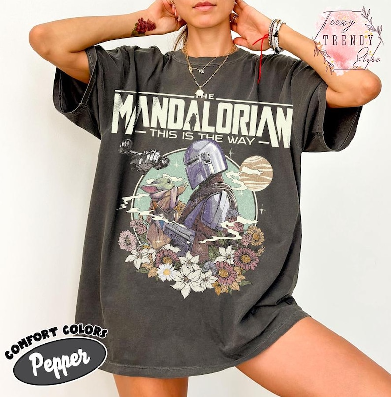 May include: Dark grey t-shirt with a graphic from The Mandalorian. The design includes the text "THE MANDALORIAN THIS IS THE WAY" with an image of the Mandalorian and Grogu surrounded by flowers. The shirt is labelled "Comfort Colors Pepper".