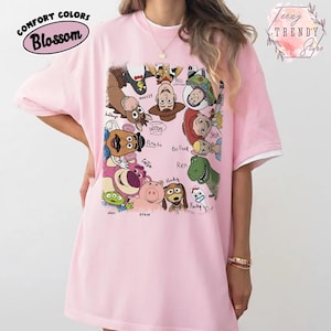 May include: A light pink t-shirt featuring a colorful graphic of characters from the animated movie Toy Story. The shirt has a white trim on the sleeves and the text "COMFORT COLORS Blossom" is printed on the upper left side.