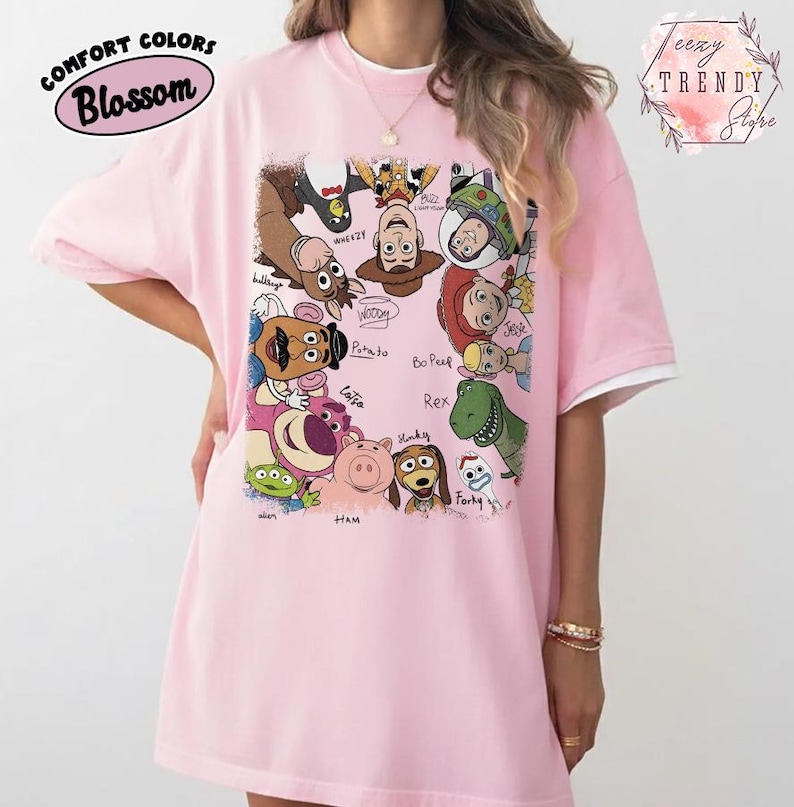 May include: A light pink t-shirt featuring a colorful graphic of characters from the animated movie Toy Story. The shirt has a white trim on the sleeves and the text "COMFORT COLORS Blossom" is printed on the upper left side.