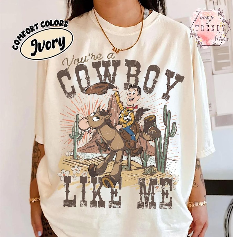 May include: An ivory t-shirt featuring a graphic of a cartoon cowboy riding a horse in a desert landscape with cacti and mountains. The text reads You're a COWBOY LIKE ME. The design uses brown, tan, and blue tones with a sunburst background.