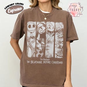 May include: An espresso brown short-sleeve t-shirt featuring a white graphic print of characters from Tim Burton's The Nightmare Before Christmas. The design includes four vertical panels displaying Jack Skellington, Sally, Oogie Boogie, and Lock, Shock, and Barrel.