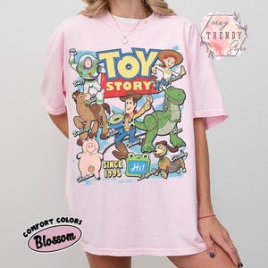 May include: A light pink t-shirt featuring a large graphic print of Toy Story characters including Buzz Lightyear, Woody, Jessie, Bullseye, Rex, Hamm, Slinky Dog, and an Alien. The text reads Toy Story, Since 1995, and Hi! with character names.