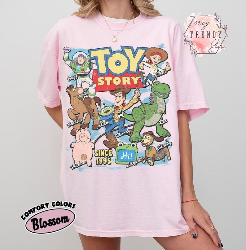 May include: A light pink t-shirt featuring a large graphic print of Toy Story characters including Buzz Lightyear, Woody, Jessie, Bullseye, Rex, Hamm, Slinky Dog, and an Alien. The text reads Toy Story, Since 1995, and Hi! with character names.