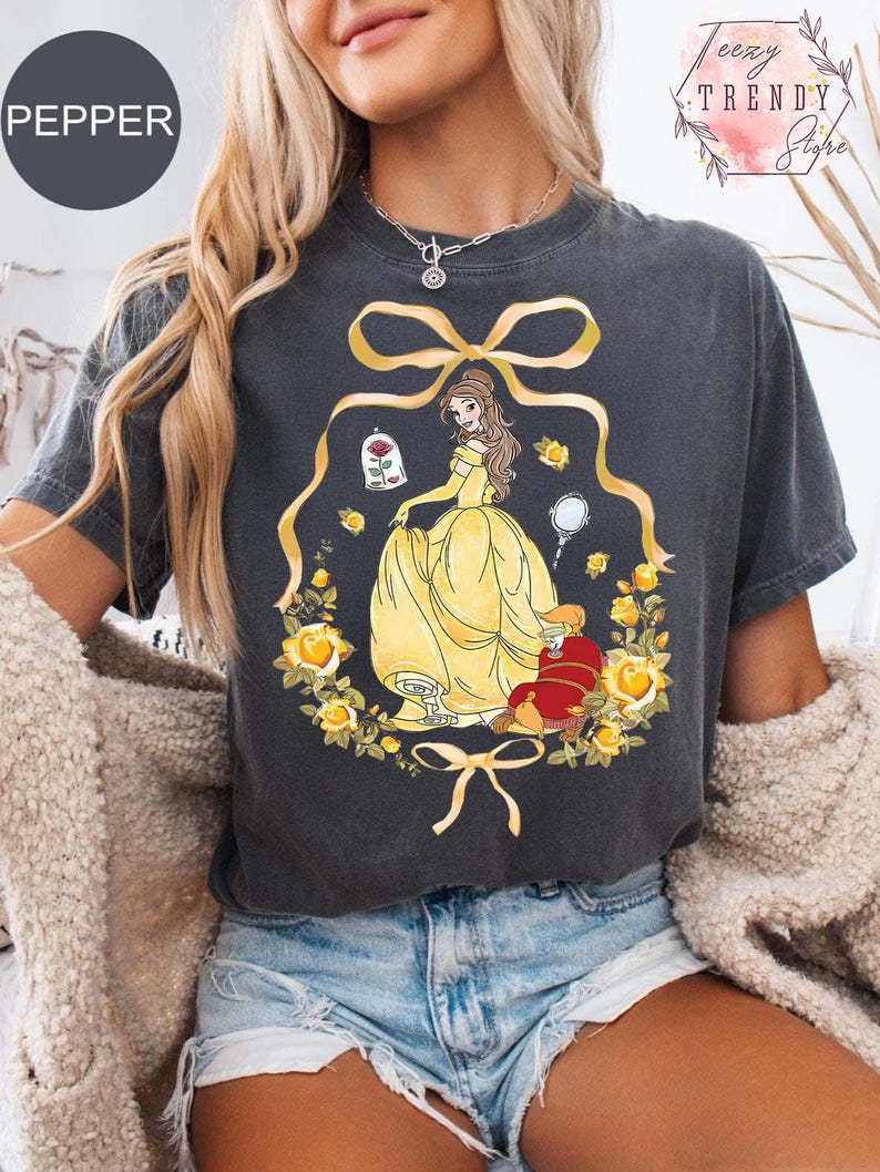 Peut inclure: T-shirt gris fonc&eacute; avec un personnage de dessin anim&eacute; en robe jaune, entour&eacute; d'une couronne florale. Le motif comprend une rose, un miroir et des n&oelig;uds. Le mot "PEPPER" est en haut &agrave; gauche.