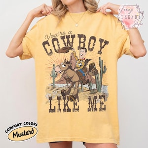 May include: A mustard yellow short-sleeve t-shirt featuring a graphic print of a cartoon cowboy riding a horse in a desert landscape with cacti. The text reads You're a COWBOY LIKE ME. The design includes a sunburst background and small flowers.