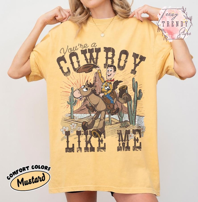 May include: A mustard yellow short-sleeve t-shirt featuring a graphic print of a cartoon cowboy riding a horse in a desert landscape with cacti. The text reads You're a COWBOY LIKE ME. The design includes a sunburst background and small flowers.