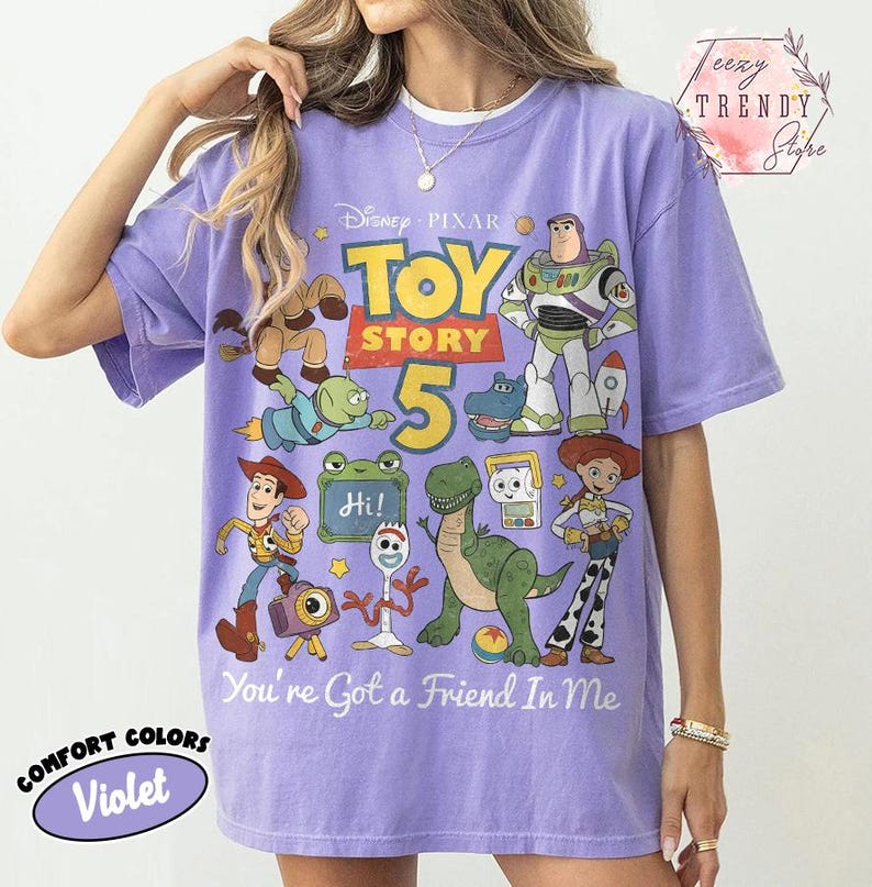 May include: A violet short-sleeve t-shirt featuring a graphic print of Disney Pixar Toy Story characters including Woody, Buzz Lightyear, Jessie, Rex, and Forky. The text reads Toy Story 5 and You've Got a Friend In Me, with a small Hi sign.