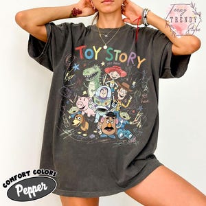 May include: Dark grey t-shirt featuring the words "Toy Story" in colorful letters, with a graphic of characters from the movie. The shirt has a vintage look with the words "Comfort Colors" and "Pepper" printed on the bottom.