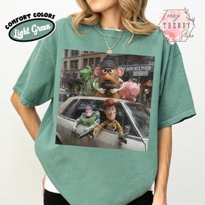 Puede incluir: Una camiseta de manga corta de color verde claro con un estampado gr&aacute;fico de cinco personajes de juguete en un coche. Los personajes incluyen un dinosaurio verde, una figura con forma de patata, un cerdo rosa, un ranger espacial y un vaquero.