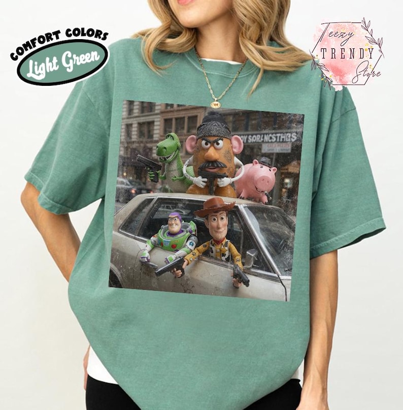 Puede incluir: Una camiseta de manga corta de color verde claro con un estampado gr&aacute;fico de cinco personajes de juguete en un coche. Los personajes incluyen un dinosaurio verde, una figura con forma de patata, un cerdo rosa, un ranger espacial y un vaquero.