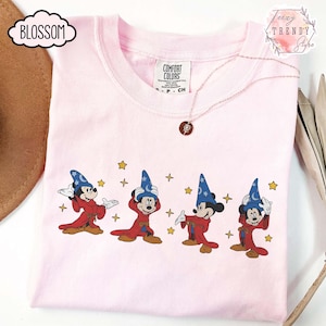 May include: A light pink t-shirt featuring a graphic of Mickey Mouse in a wizard costume with a blue hat and red robe. The design includes yellow stars. A silver necklace with a pendant is on the shirt.