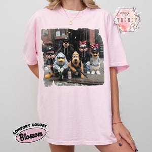 May include: A light pink t-shirt featuring a graphic print of cartoon characters wearing streetwear, including hoodies, sunglasses, and baseball caps. The characters are positioned in an urban setting. The shirt has a crew neckline and short sleeves.