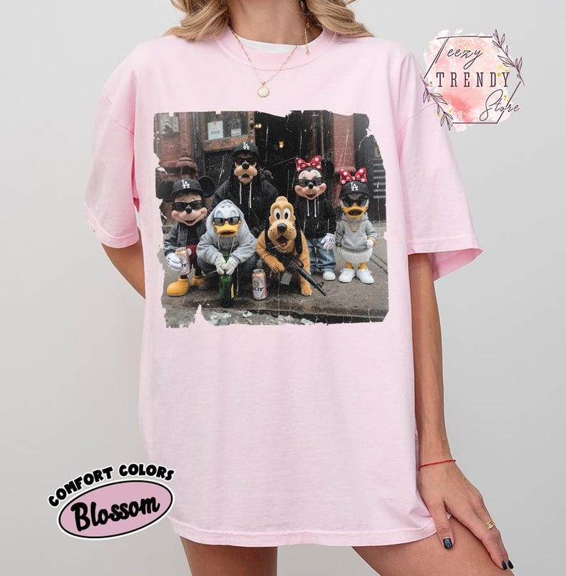 May include: A light pink t-shirt featuring a graphic print of cartoon characters wearing streetwear, including hoodies, sunglasses, and baseball caps. The characters are positioned in an urban setting. The shirt has a crew neckline and short sleeves.