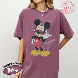 May include: Berry-colored Comfort Colors t-shirt featuring a graphic of Mickey Mouse wearing pink sunglasses. The cartoon character is in a classic pose with a white glove and red shorts. The words "Mickey Mouse" are written in cursive.