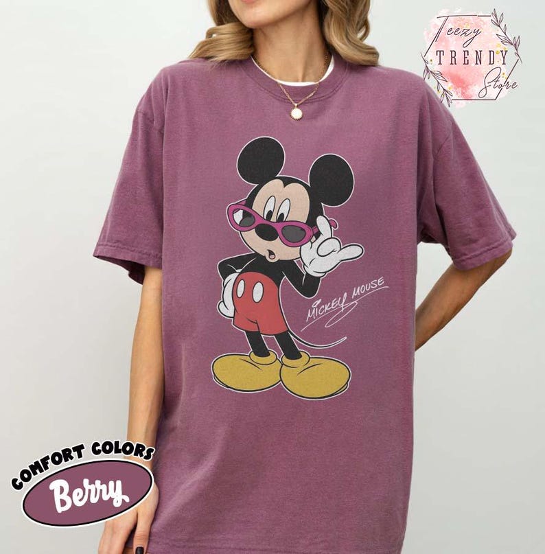 May include: Berry-colored Comfort Colors t-shirt featuring a graphic of Mickey Mouse wearing pink sunglasses. The cartoon character is in a classic pose with a white glove and red shorts. The words "Mickey Mouse" are written in cursive.
