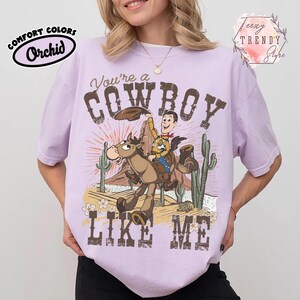 May include: A light purple short-sleeve t-shirt featuring a graphic of a cowboy riding a horse in a desert landscape with cacti and mountains. The text reads You're a COWBOY LIKE ME. The shirt includes a Comfort Colors Orchid label and a Teezy Trendy Store logo.