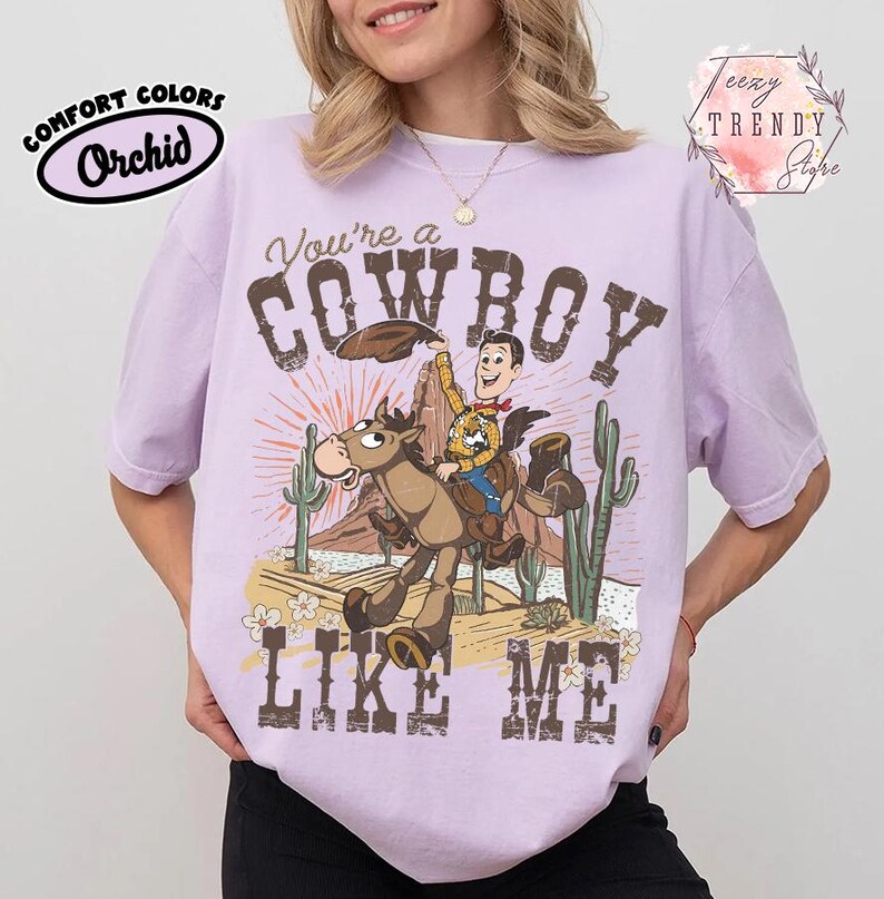 May include: A light purple short-sleeve t-shirt featuring a graphic of a cowboy riding a horse in a desert landscape with cacti and mountains. The text reads You're a COWBOY LIKE ME. The shirt includes a Comfort Colors Orchid label and a Teezy Trendy Store logo.