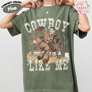 May include: A moss green short-sleeve t-shirt featuring a graphic print of a cartoon cowboy riding a horse in a desert landscape. The text "You're a COWBOY LIKE ME" is printed in a distressed white font around the central illustration.