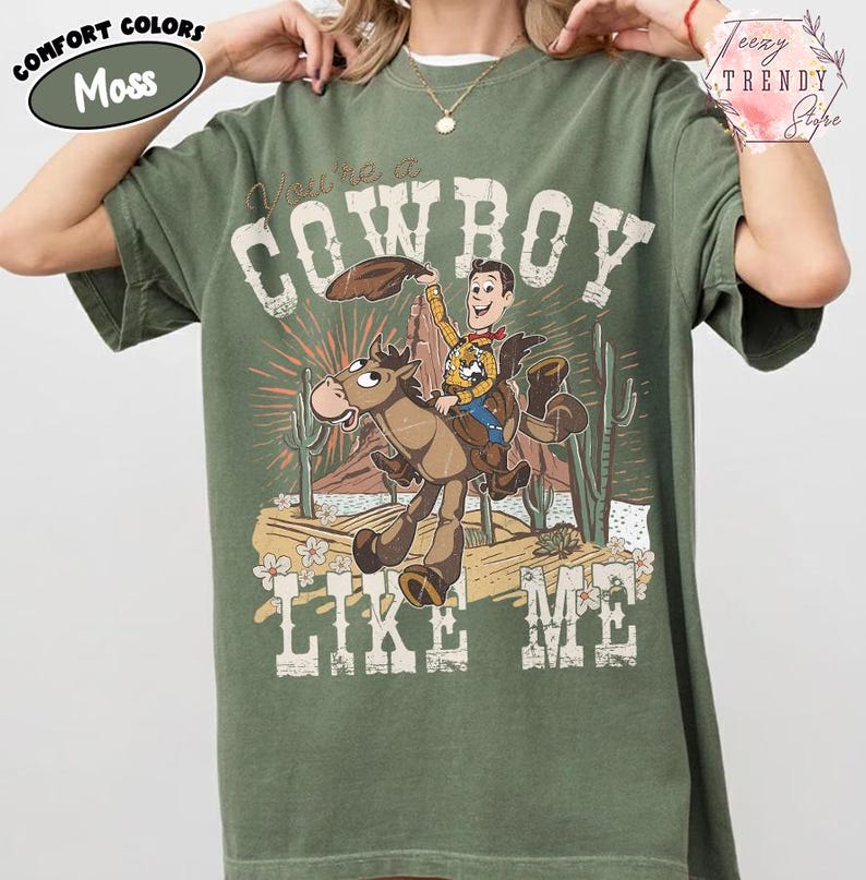 May include: A moss green short-sleeve t-shirt featuring a graphic print of a cartoon cowboy riding a horse in a desert landscape. The text "You're a COWBOY LIKE ME" is printed in a distressed white font around the central illustration.
