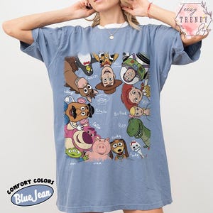 May include: Blue jean colored t-shirt featuring a graphic of various Toy Story characters. The shirt has a crew neck and short sleeves. The words "COMFORT COLORS" and "Blue Jean" are printed on the bottom of the shirt.