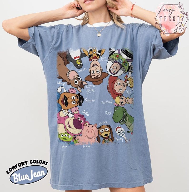 May include: Blue jean colored t-shirt featuring a graphic of various Toy Story characters. The shirt has a crew neck and short sleeves. The words "COMFORT COLORS" and "Blue Jean" are printed on the bottom of the shirt.